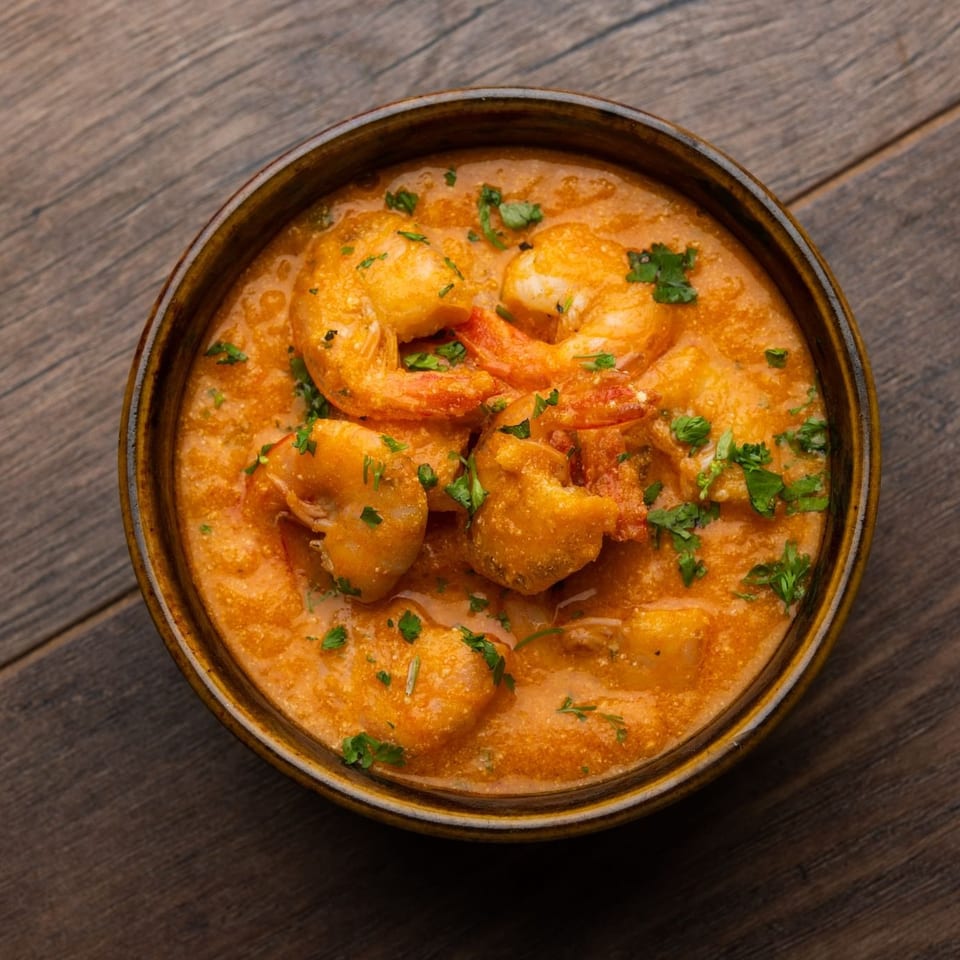 A vibrant orange shrimp curry, garnished with fresh green herbs, served in a brown bowl on wood.
