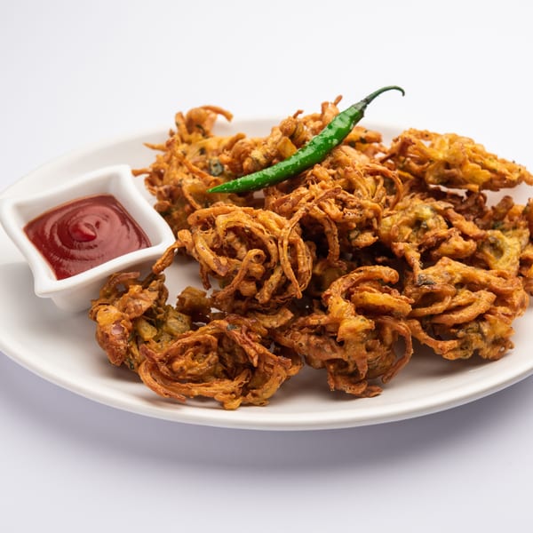 Crispy golden onion pakora with a green chili and a side of red ketchup on a white plate.