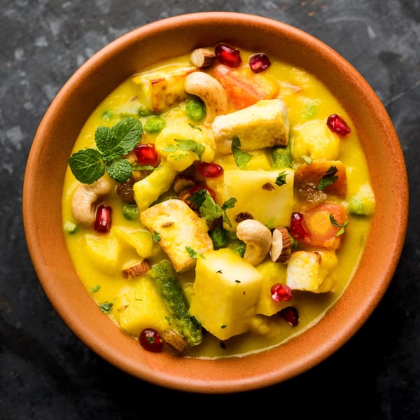 A vibrant yellow curry with paneer, vegetables, cashews, and pomegranate seeds in a terracotta bowl.