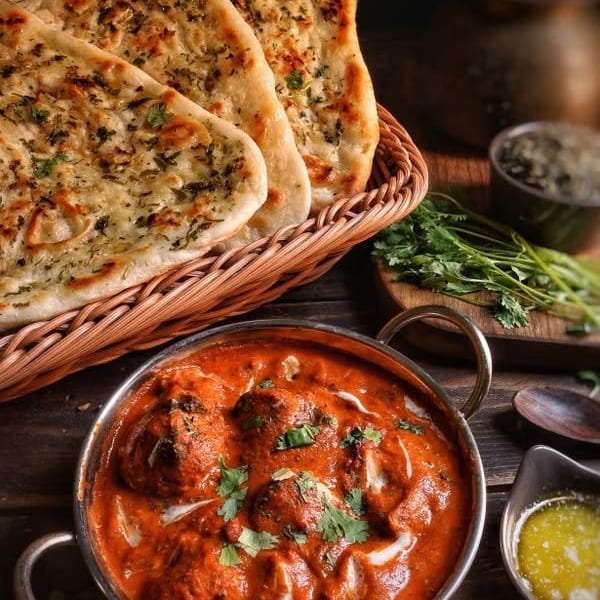 Indian butter chicken in a metal bowl with garlic naan bread in a basket.