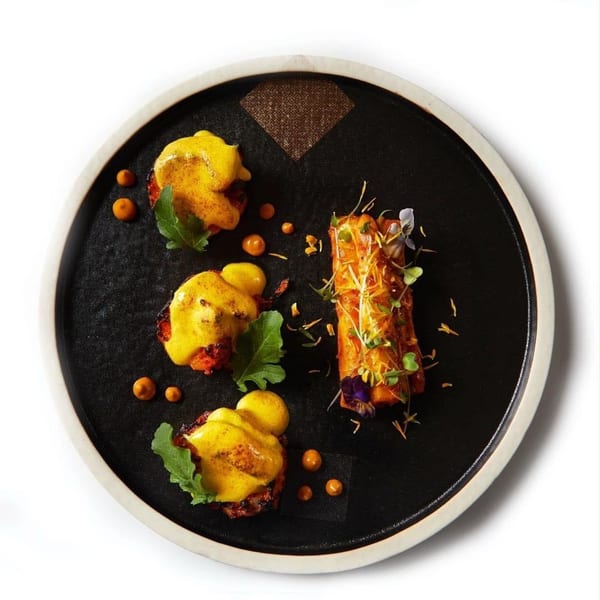 Artfully plated dish with three yellow-sauced appetizers and a garnished roll on a black plate.