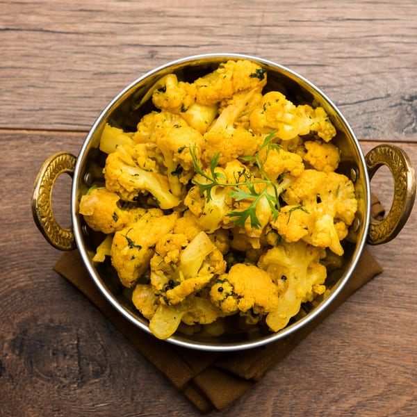 A brass bowl filled with yellow spiced cauliflower florets, garnished with fresh cilantro, on a wooden table.